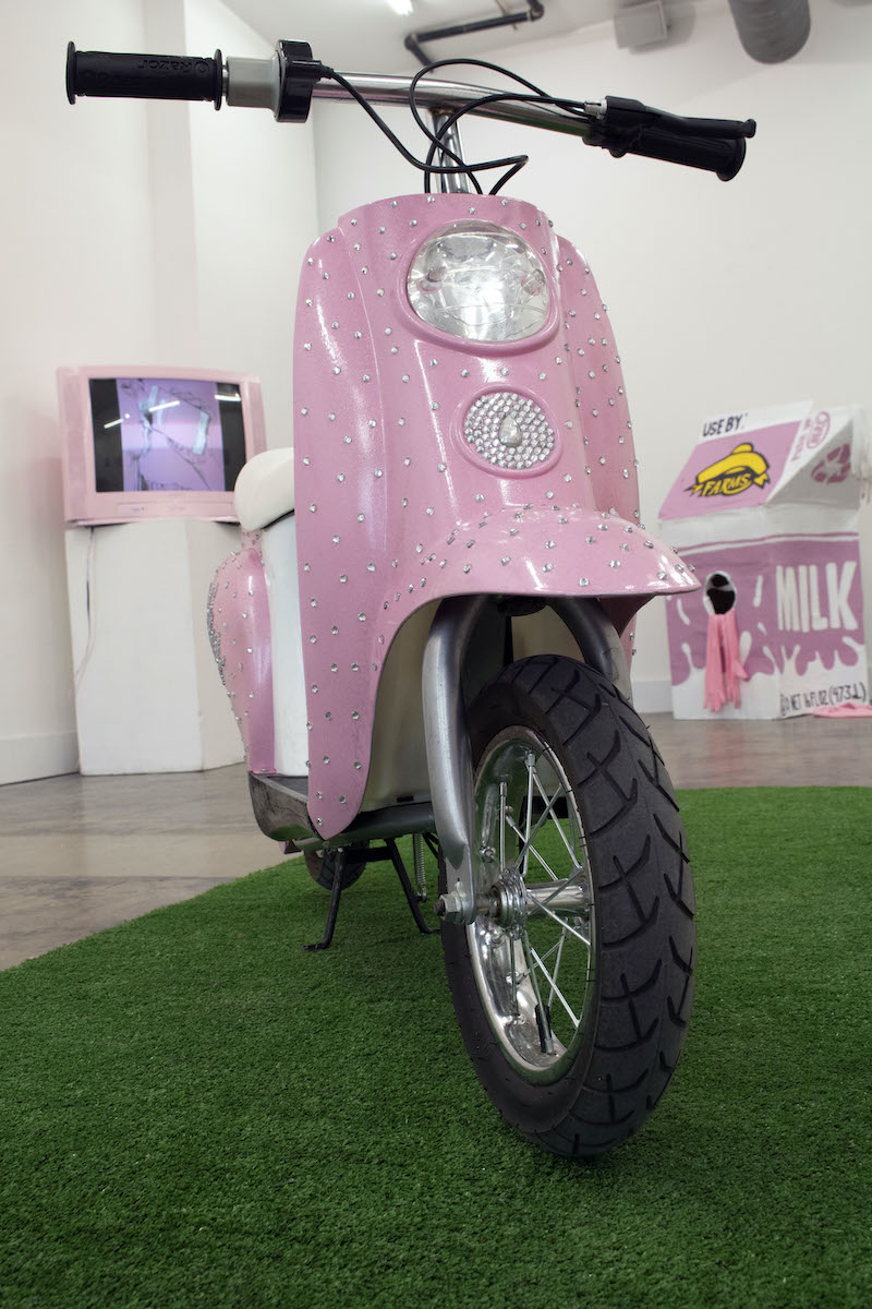 A pink moped with the pink tv and lifesize milk carton in the background