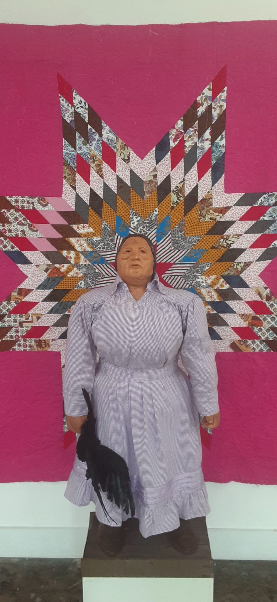 A doll-like figure of a woman in  lavender clothing in front of a pink quilt
