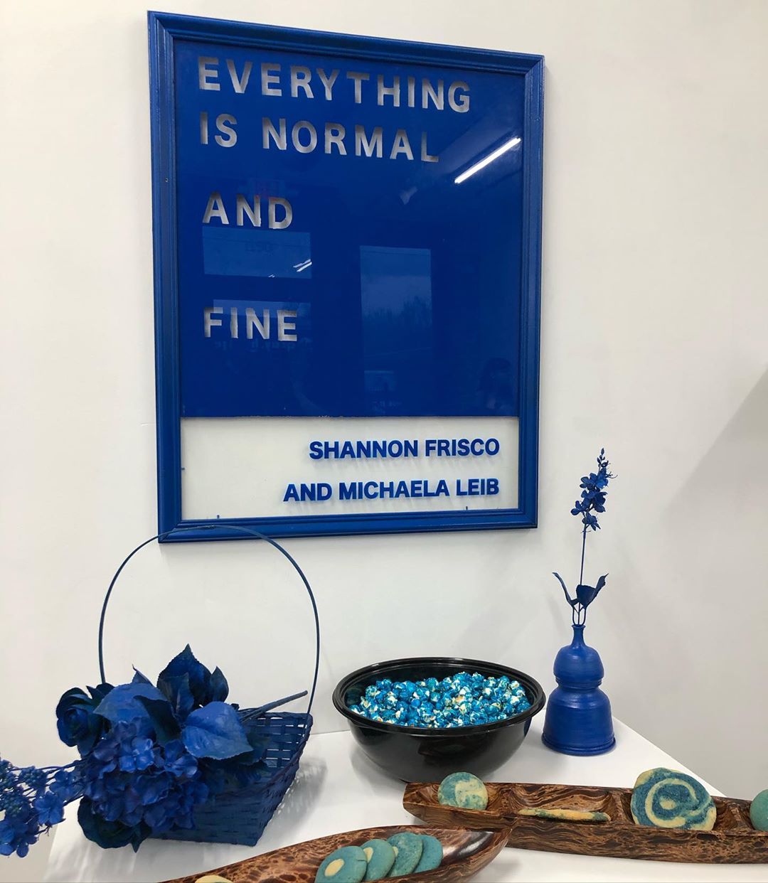 Blue and white sign reading Everything is Normal and Fine, Shannon Frisco and Michaela Leib, assorted blue and green items on the table beneath