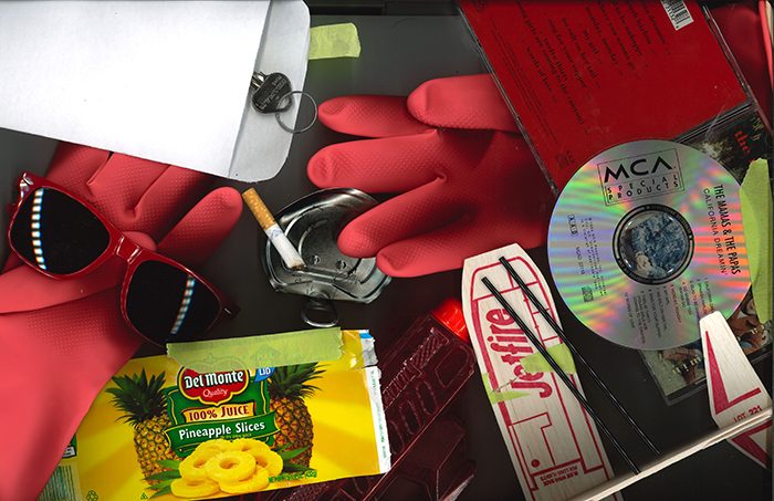 A photo of an assortment of objects including a cd, red gloves, a cigarette, a key in an envelope, and a can of pineapple slices