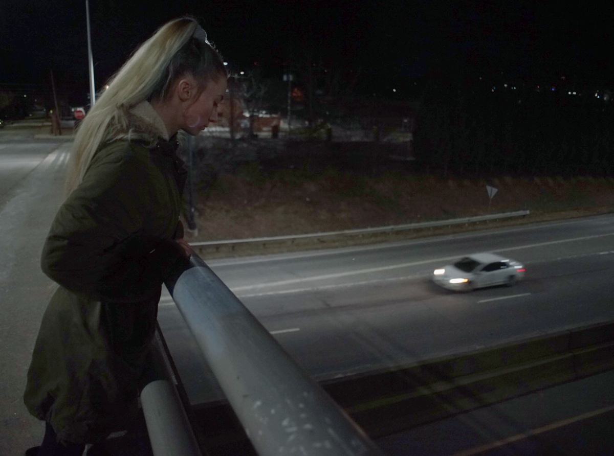 A photo of a young blonde woman at night looking over a highway overpass, a white car passing underneath