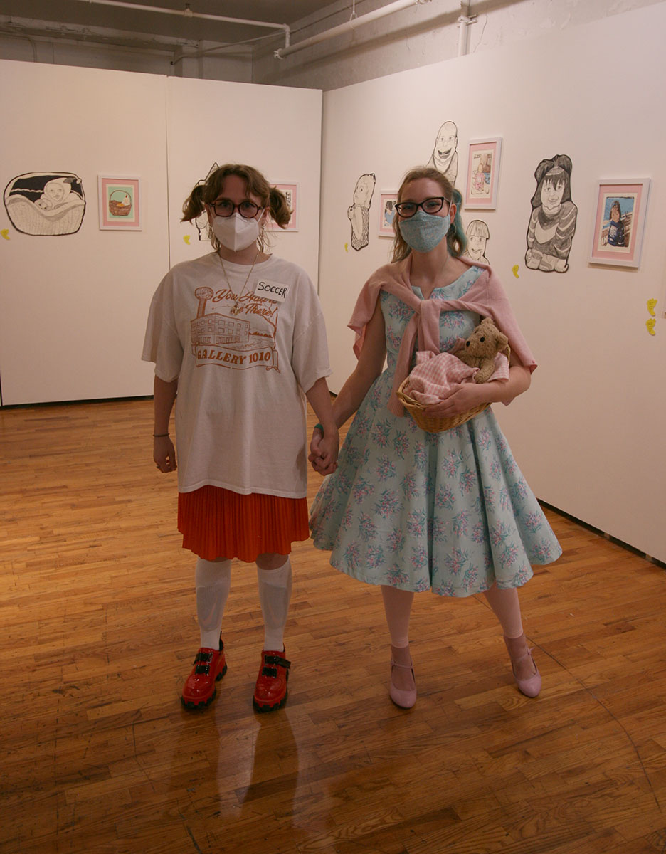 Two masked woman holding hands, one of them holding a swaddled teddy bear while surrounded by the "Baby Show" installation