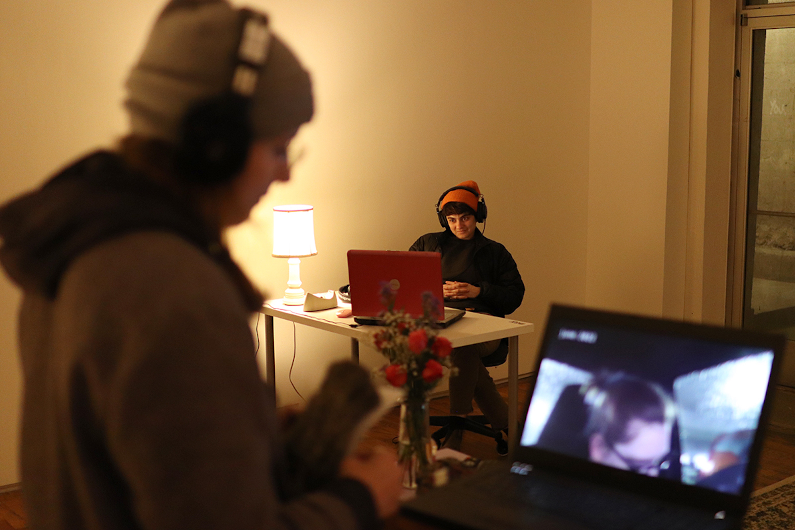 One person watches a video on a laptop while a person in the background watches on another laptop with headphones on