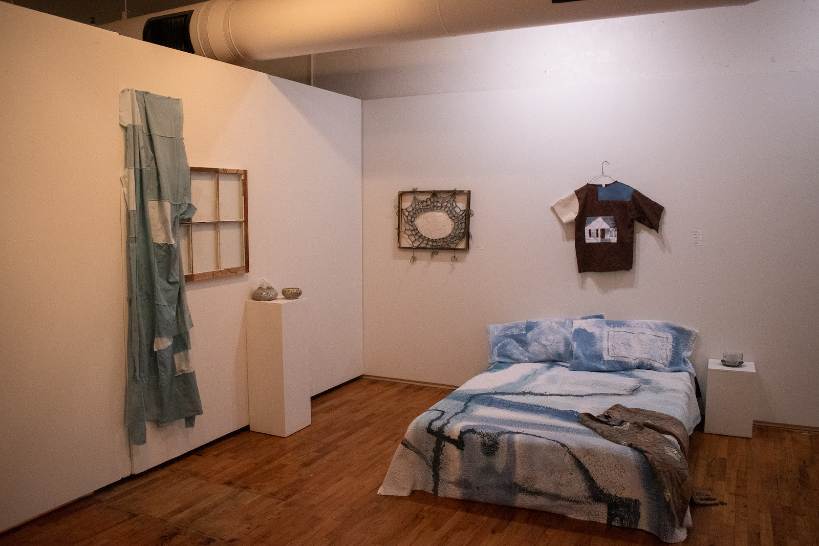 A bedroom set up in an art gallery, including a bed, a glass-less window frame, and a shirt on a hanger on the wall