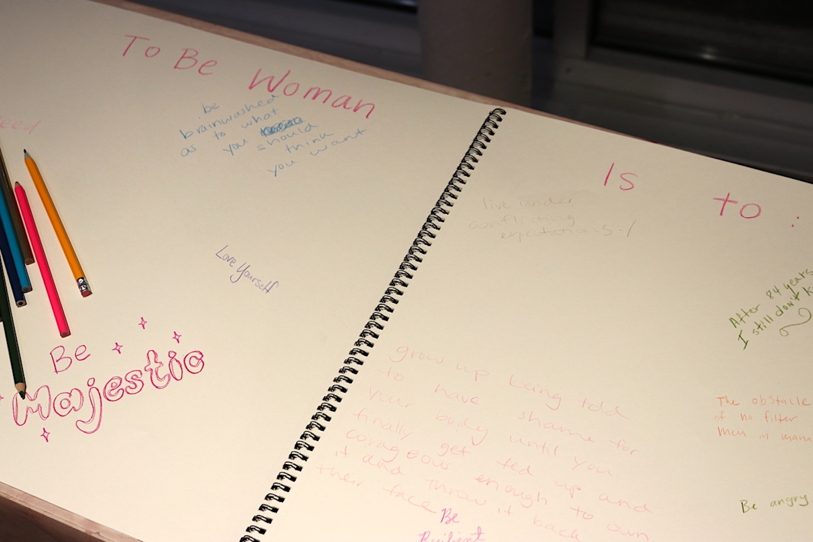 A notebook with the words "To Be Woman Is To:" at the top and various messages written including "Love Yourself" and "Be Majestic"