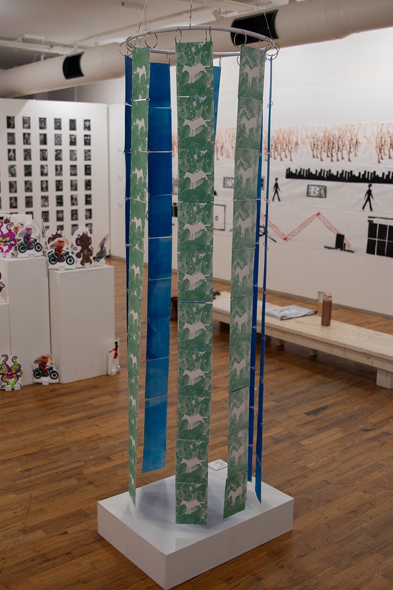 An art gallery installation featuring colorful, postcard-sized illustrated sheets suspended from the ceiling