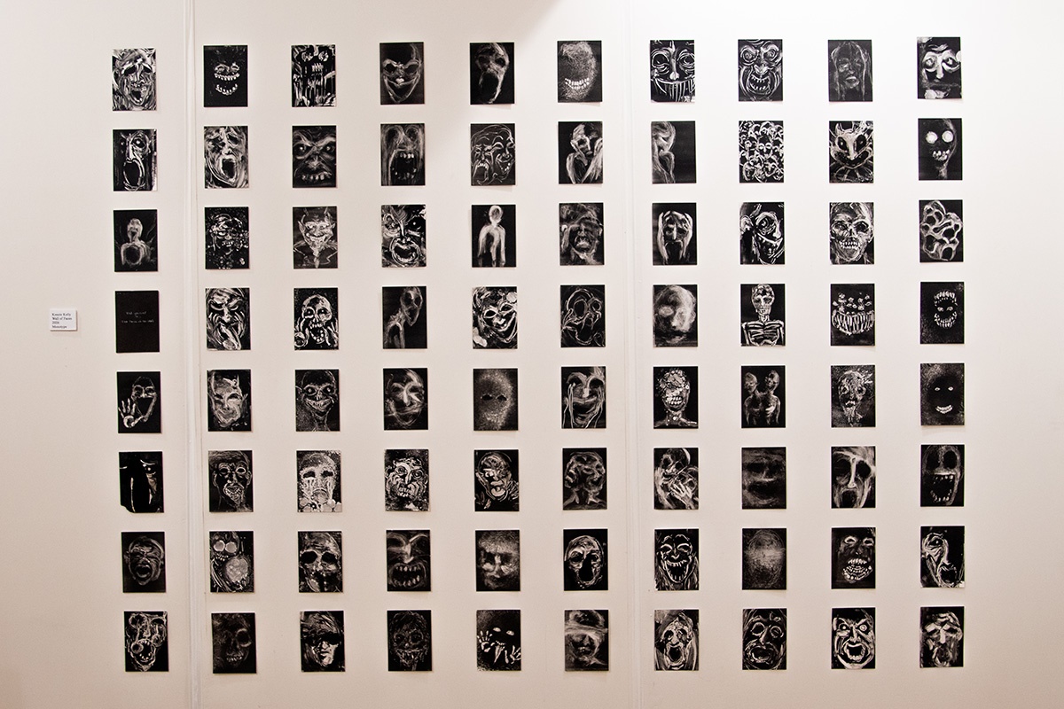 An art gallery wall displaying dozens of small black-and-white artworks arranged in a regular grid, depicting abstract and distorted faces and figures.