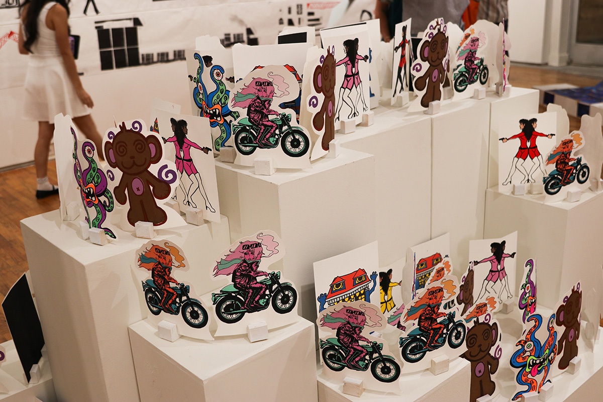 An art gallery installation featuring colorful illustrated cutout figures, including monkeys, motorcycles, women, and abstract creatures, arranged on white geometric pedestals.