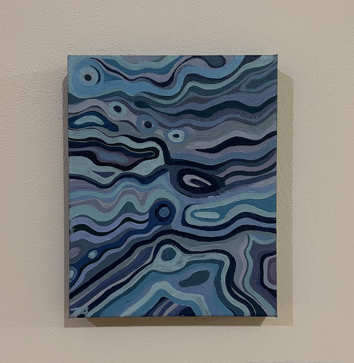 Painting titled 'Nevada Sky' by Nolan Steil, mounted on a light-colored wall. The abstract post-impressionist composition features flowing, marbled bands of blue, gray, and purple in varying shades, creating a swirling, map-like or aerial pattern. Small circular and oval shapes in lighter and darker tones are scattered throughout, resembling clouds, pools, or celestial forms embedded within the wavy lines