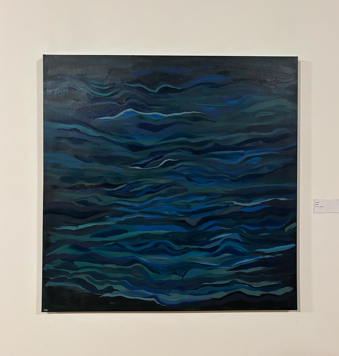 Painting titled 'Pacific' by Nolan Steil, mounted on a light-colored wall with a small text label to its right. The square canvas depicts an abstract, wave-like composition in deep blues, teals, and blacks, resembling rippling ocean water viewed from above. Undulating horizontal bands of varying shades create a sense of depth and movement across the surface, with a small artist's signature in the bottom left corner