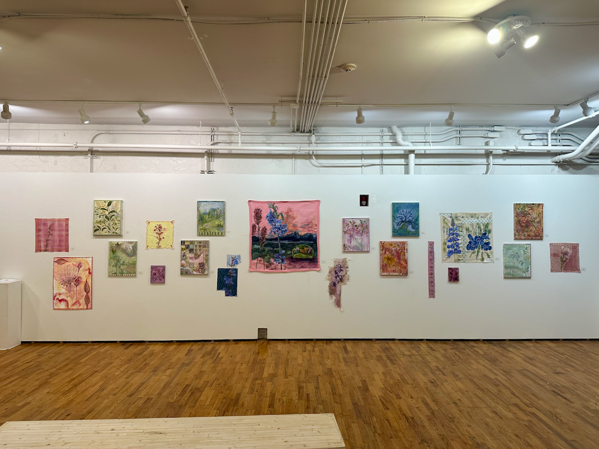 Gallery installation view of a long white wall densely arranged with numerous small to medium-sized artworks featuring botanical and floral motifs in a mix of media, including paintings, textiles, and mixed-media pieces. Colors range from soft pinks, greens, and yellows to blues and purples. A larger pink-toned painting depicting a landscape with tall blue flowers and a distant tree line hangs near the center.