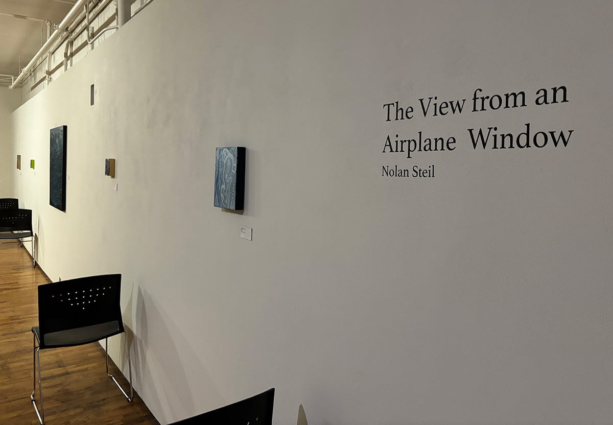 Gallery installation view of a long white wall receding into the distance, displaying the exhibition title 'The View from an Airplane Window' by Nolan Steil in black text on the right. Several small paintings are mounted along the wall, including abstract swirling blue and white compositions and a few smaller dark and yellow-toned pieces, each with a small wall label.