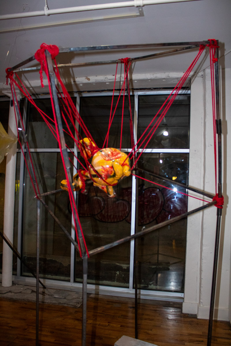 Yellow ceramic sculpture suspended in a metal frame with red rope.