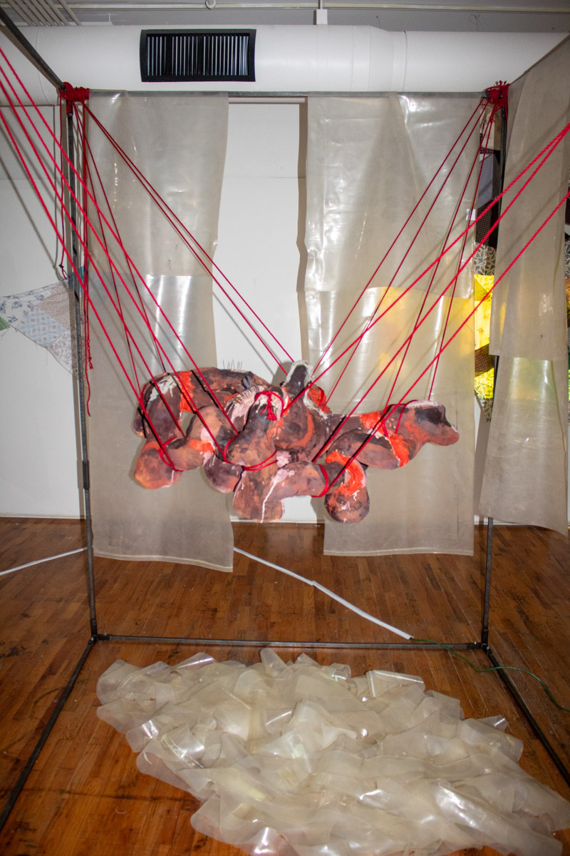 Ceramic object suspended with red rope in a metal frame with a plastic curtain. Scraps of plastic in a pile underneath the ceramic pieces.