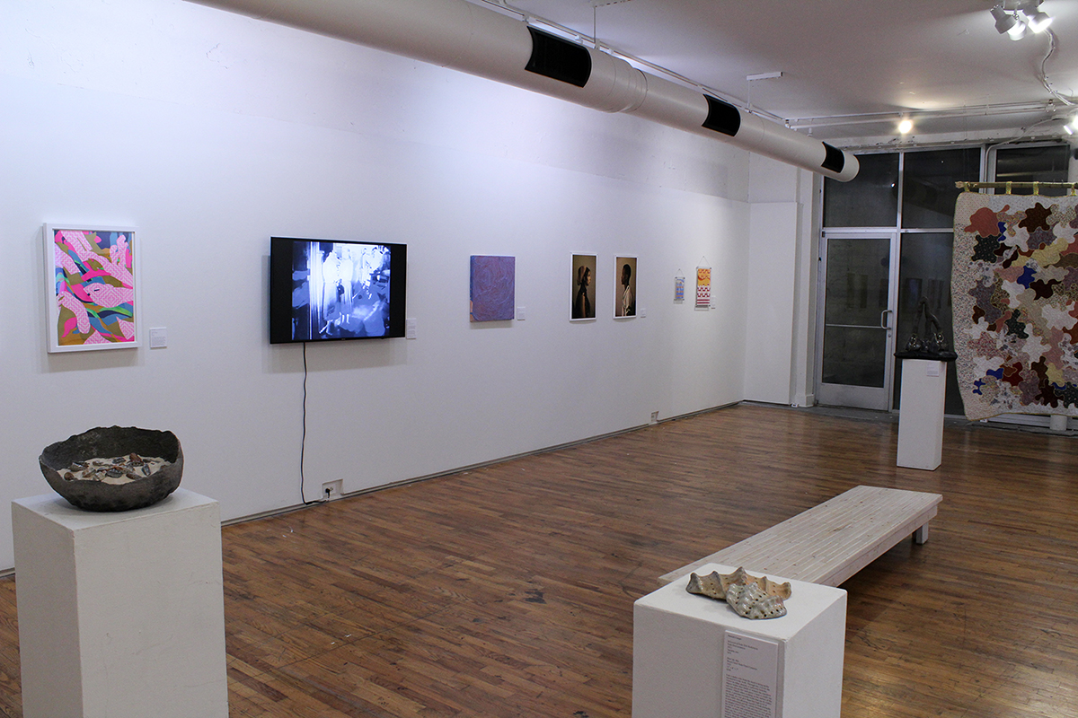 Gallery with framed prints, a video monitor, photographs, and a patchwork quilt on the walls, and ceramic and sculptural objects on white pedestals.